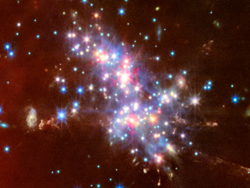     A imagem mostra um denso aglomerado de galáxias de fundo e estruturas nebulosas vermelhas na área. As cores da imagem correspondem aos diferentes filtros das câmeras MIRI e NIRCam 